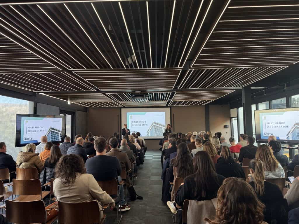 Public assis dans une salle au plafond à lattes, face à trois écrans de présentation