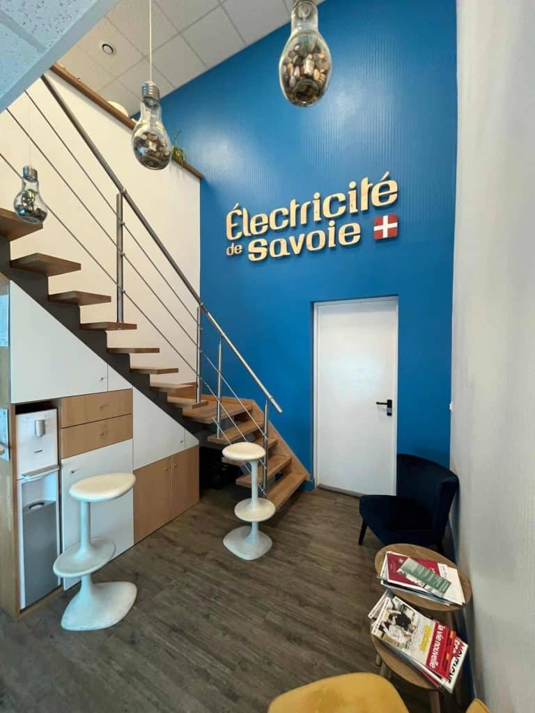 Accueil des bureaux d’Électricité de Savoie à Saint-Julien-Mont-Denis, réaménagés par INSIDE : escalier en bois et logo sur un mur bleu