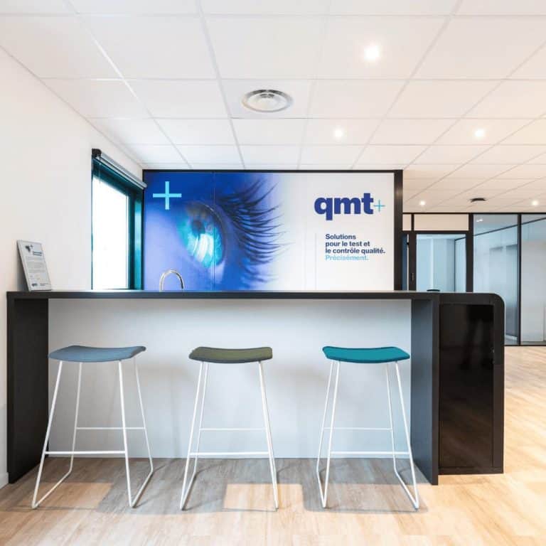 Banque d’accueil de QMT France à Alpespace, meublée par WOM : tabourets hauts et logo de l’entreprise