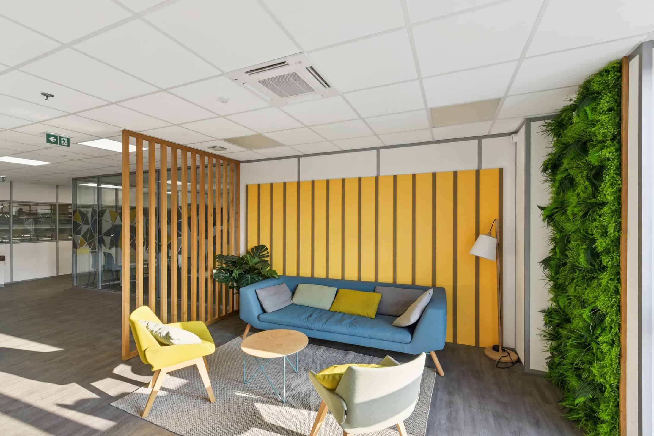Espace détente meublé par WOM chez Stryker, en Isère : canapé bleu, fauteuils jaunes, claustra en bois et mur végétal